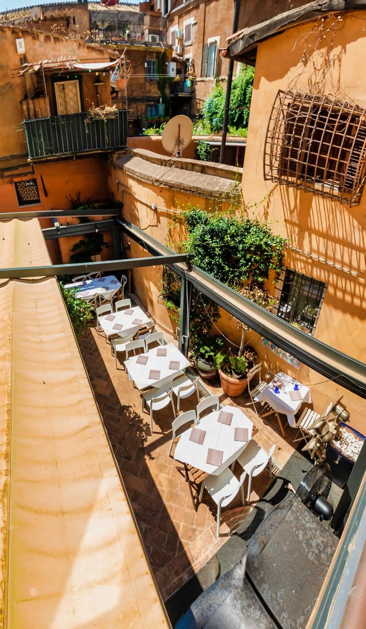 A terrace with white-clothed tables and chairs sits in a narrow courtyard between ochre-walled buildings, seen from above.