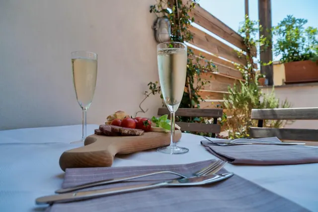 An outdoor terrace table is set with two flutes of sparkling wine, a wooden board of bread, tomatoes and cured meat, and cutlery on folded napkins.