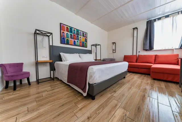 A hotel bedroom with a large bed with a maroon throw, a red sofa, a purple armchair and a colourful arched artwork on the wall.