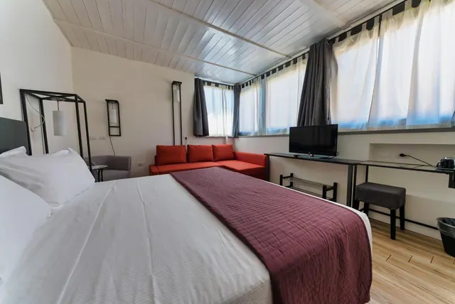 A bedroom with a double bed under a burgundy throw, a red sofa, a grey armchair, and a wall-mounted television on a desk beside large windows with white curtains.