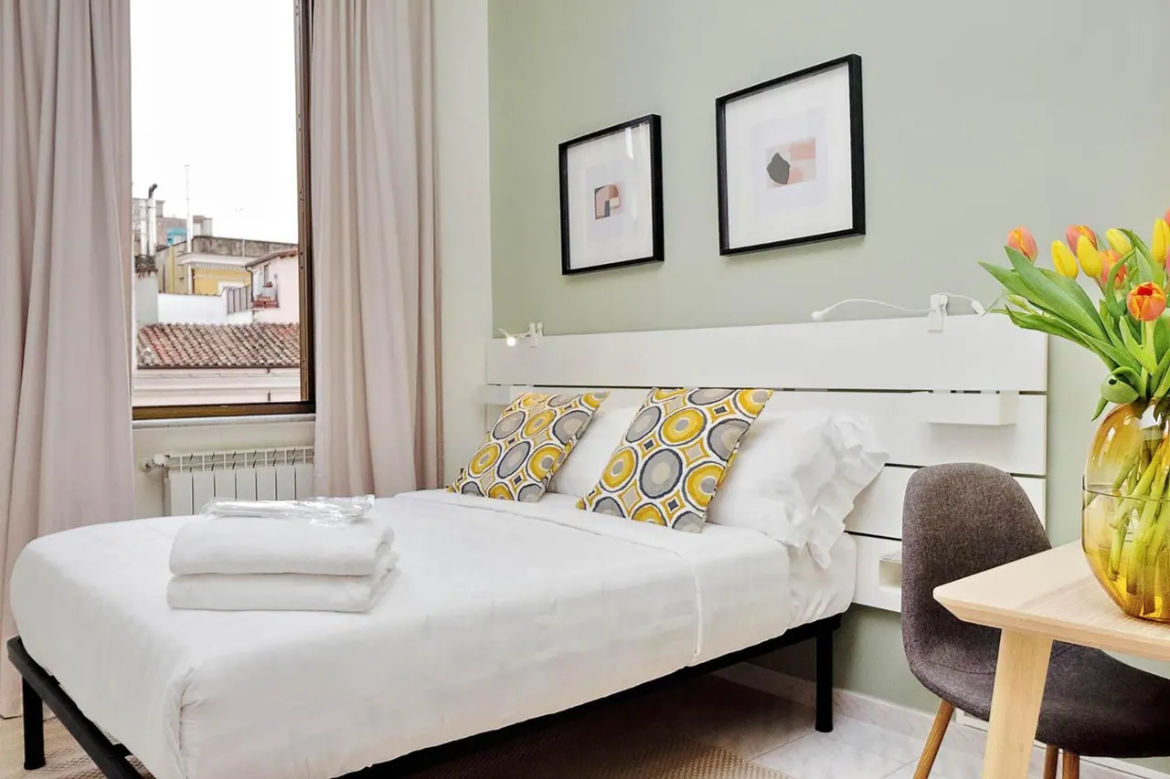 A double bed with white linens, yellow patterned cushions and folded towels sits beside a window looking over rooftops, with a small table and tulips at right.