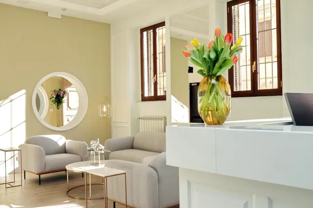 A bright lobby with grey armchairs, small white side tables, a round wall mirror and a white reception desk holding a vase of tulips.