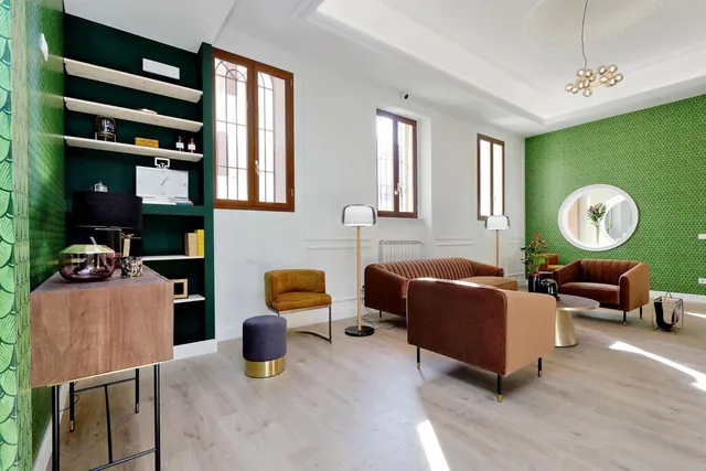 A lounge area with green patterned wallpaper, brown and tan sofas and armchairs, floor lamps, and built-in dark green shelving.