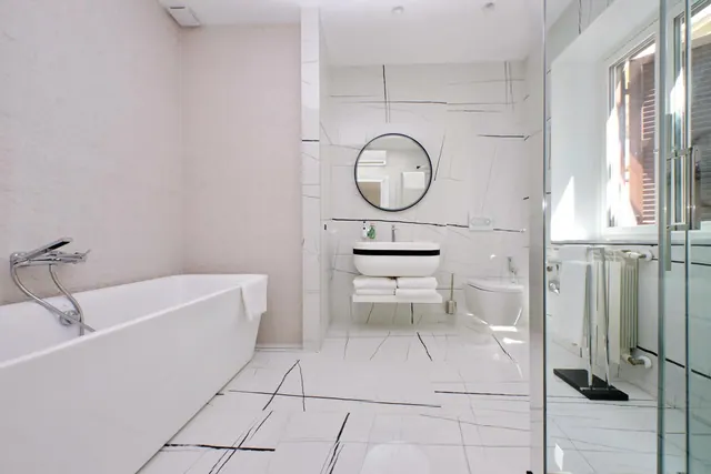 A white marble-look bathroom with a freestanding bathtub, a round wall mirror, a floating vanity with a vessel sink and stacked towels, and a glass shower enclosure beside a window.