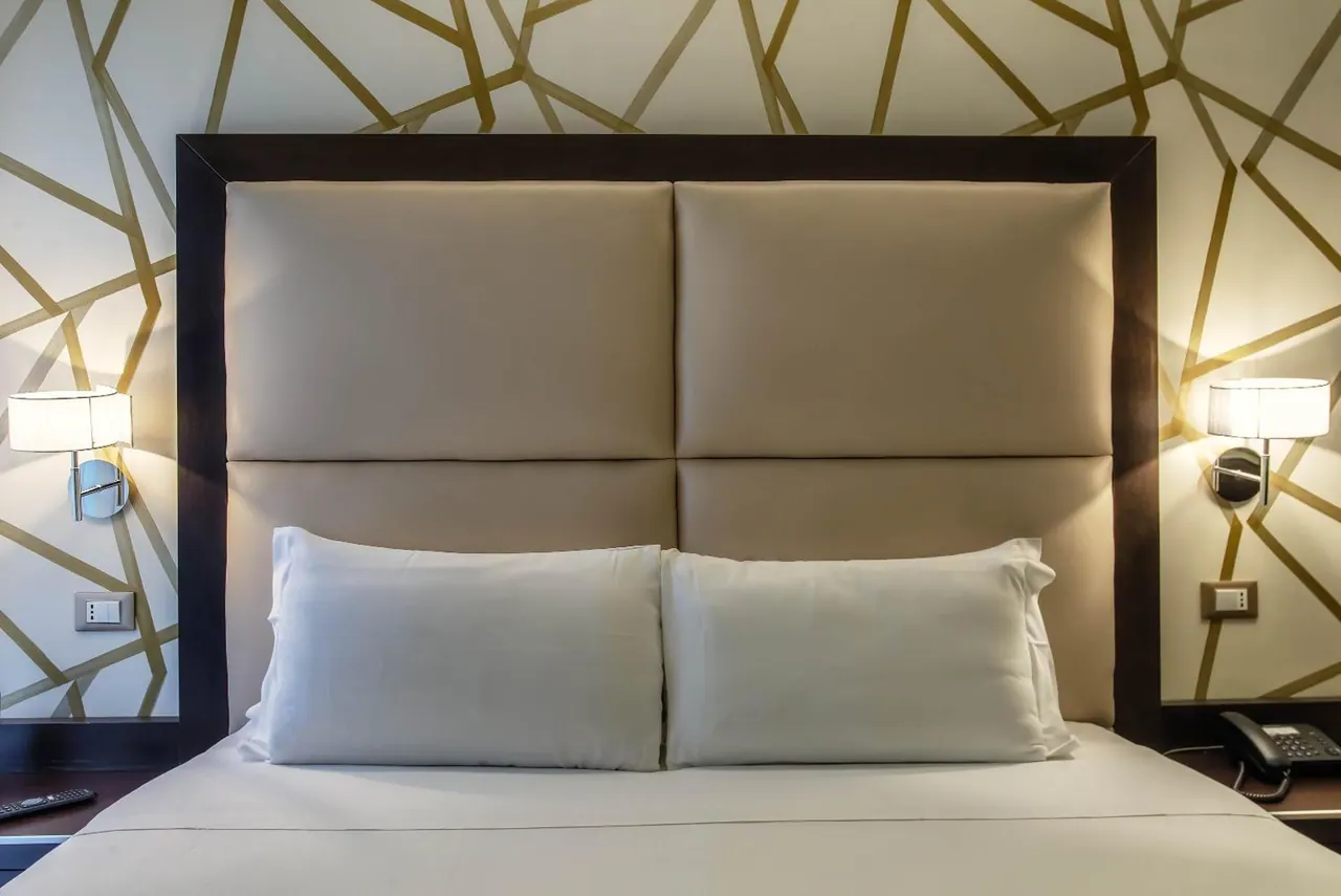 A made-up double bed with two white pillows sits against an upholstered headboard, with wall lamps on either side and a telephone on the nightstand.