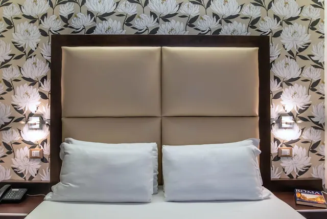 A made bed with a tall padded tan headboard, white pillows, floral wallpaper, and a telephone and a Roma guidebook on the nightstands.