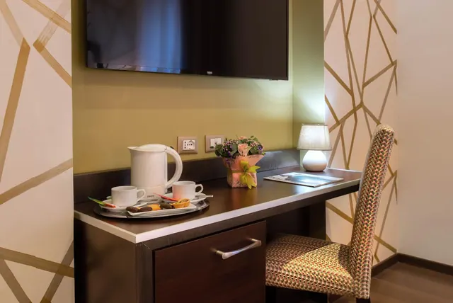 A wooden desk with a wall-mounted television above it holds a tray with an electric kettle, two cups, snacks, a flower vase and a table lamp, with an upholstered chair beside it against a geometric gold-patterned wall.