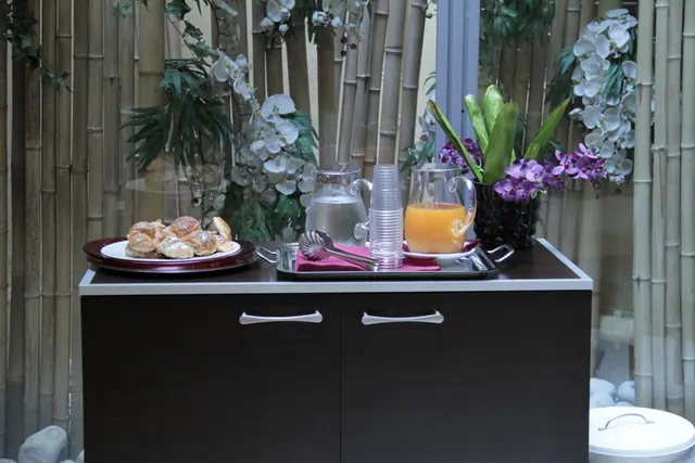 A breakfast serving station holds pastries on a plate, a water pitcher, stacked plastic cups, a pitcher of orange juice and tongs, with bamboo and white orchids behind it.