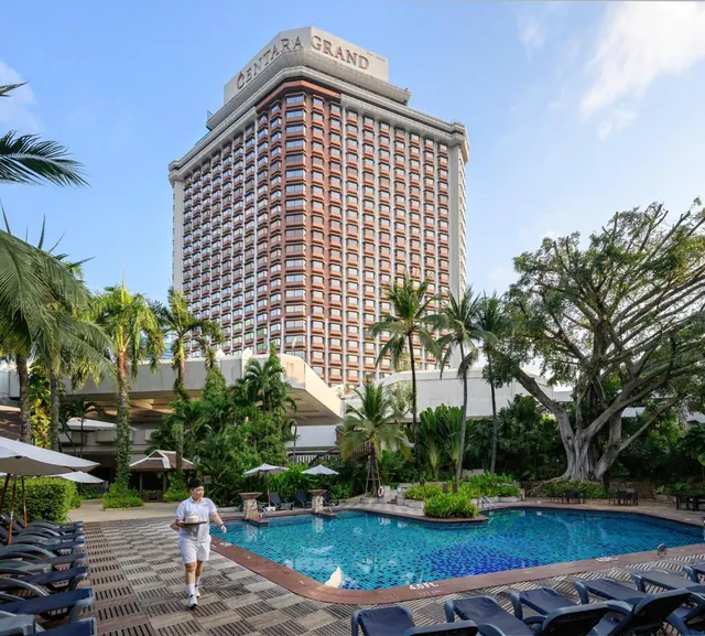 A staff member carries a tray past a large swimming pool with sun loungers, with the Centara Grand hotel tower rising behind.