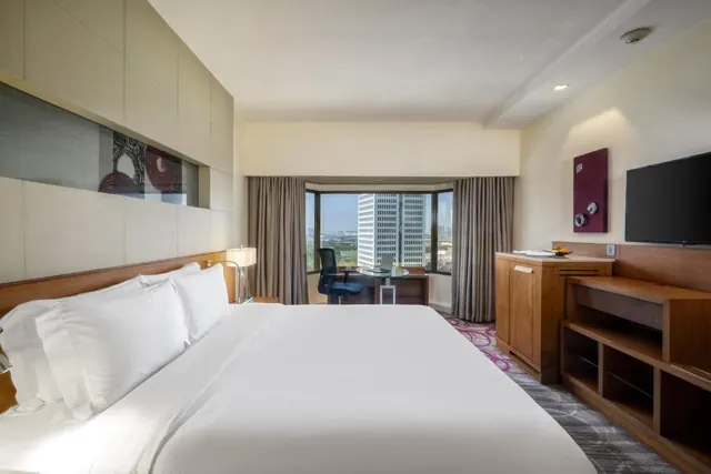 A bedroom with a made king bed, a work desk by a window looking out over a city skyline, a wooden cabinet and a wall-mounted television.