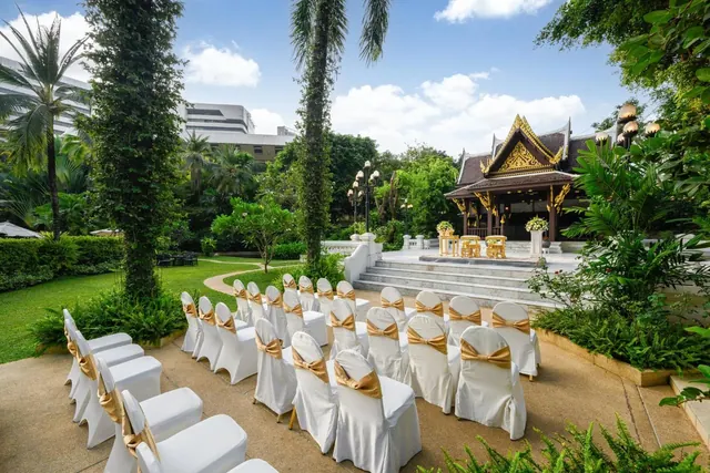 Rows of white chairs with gold sashes are set out facing a Thai-style pavilion with golden gables in a garden.