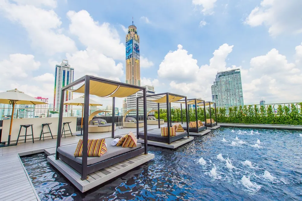 A rooftop pool with cabana daybeds on platforms in the water, with city towers and a tall skyscraper in the background.