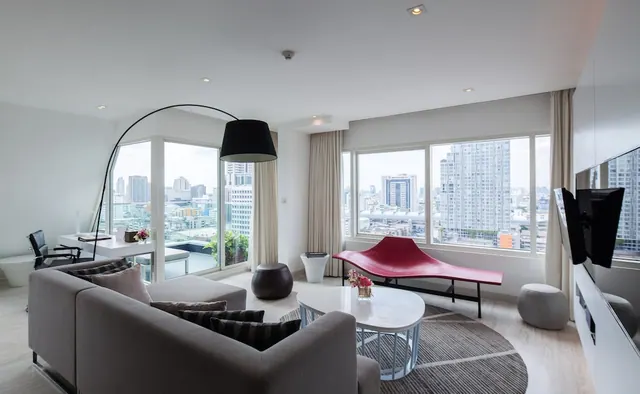 A bright living area with a grey sofa, a red chaise, an arc floor lamp and floor-to-ceiling windows overlooking high-rise buildings.