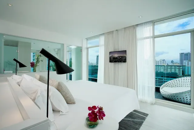 A white bedroom with a large bed, a wall-mounted television and floor-to-ceiling windows opening onto a balcony with a round wicker chair and a view of high-rise buildings.