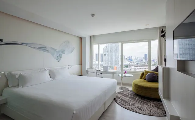 A bedroom with a made white bed, a desk and chair by the window, a round olive-green seating piece, and a city skyline visible through the windows.