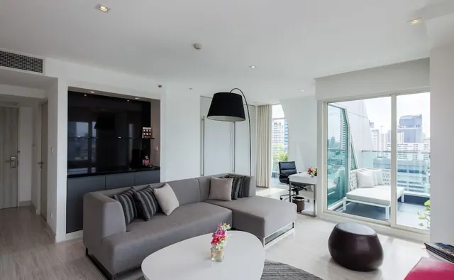 A living area with a grey sectional sofa, a wall-mounted television, and a glass sliding door opening onto a balcony with a white daybed and a view of high-rise buildings.