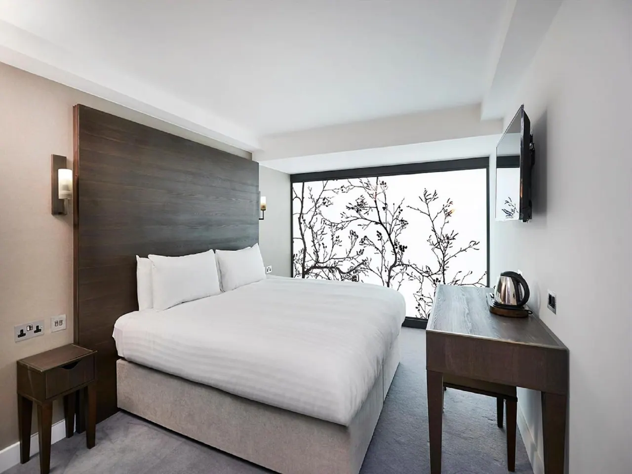 A double bed with white bedding and a dark wood headboard sits beside a wooden desk with a kettle and a wall-mounted television, with a large window behind the bed.