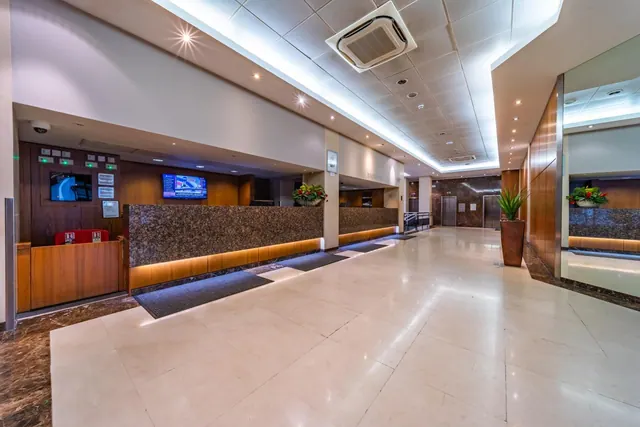 A hotel lobby with a dark marble reception counter, wall-mounted TV screens, potted plants, a large mirror on the right and lift doors at the far end.