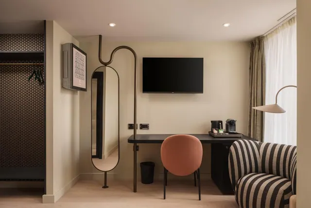 A hotel room with a wall-mounted television above a black desk and orange chair, a kettle and coffee machine on a tray, a striped armchair in the foreground, and an open wardrobe on the left.