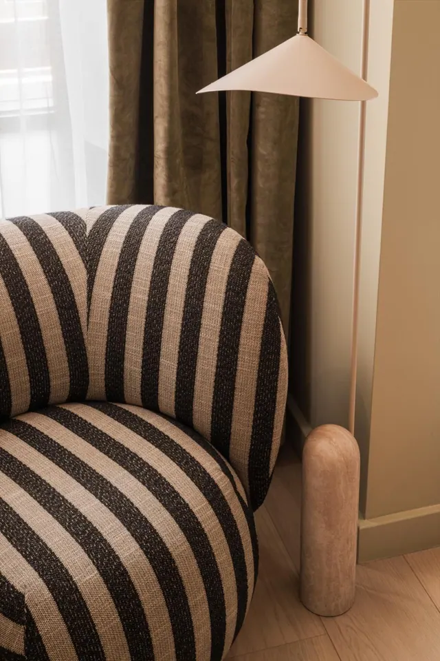 A striped armchair sits beside a olive velvet curtain and a cone-shaped lamp on a stone pillar base, with sheer curtains at the window on the left.