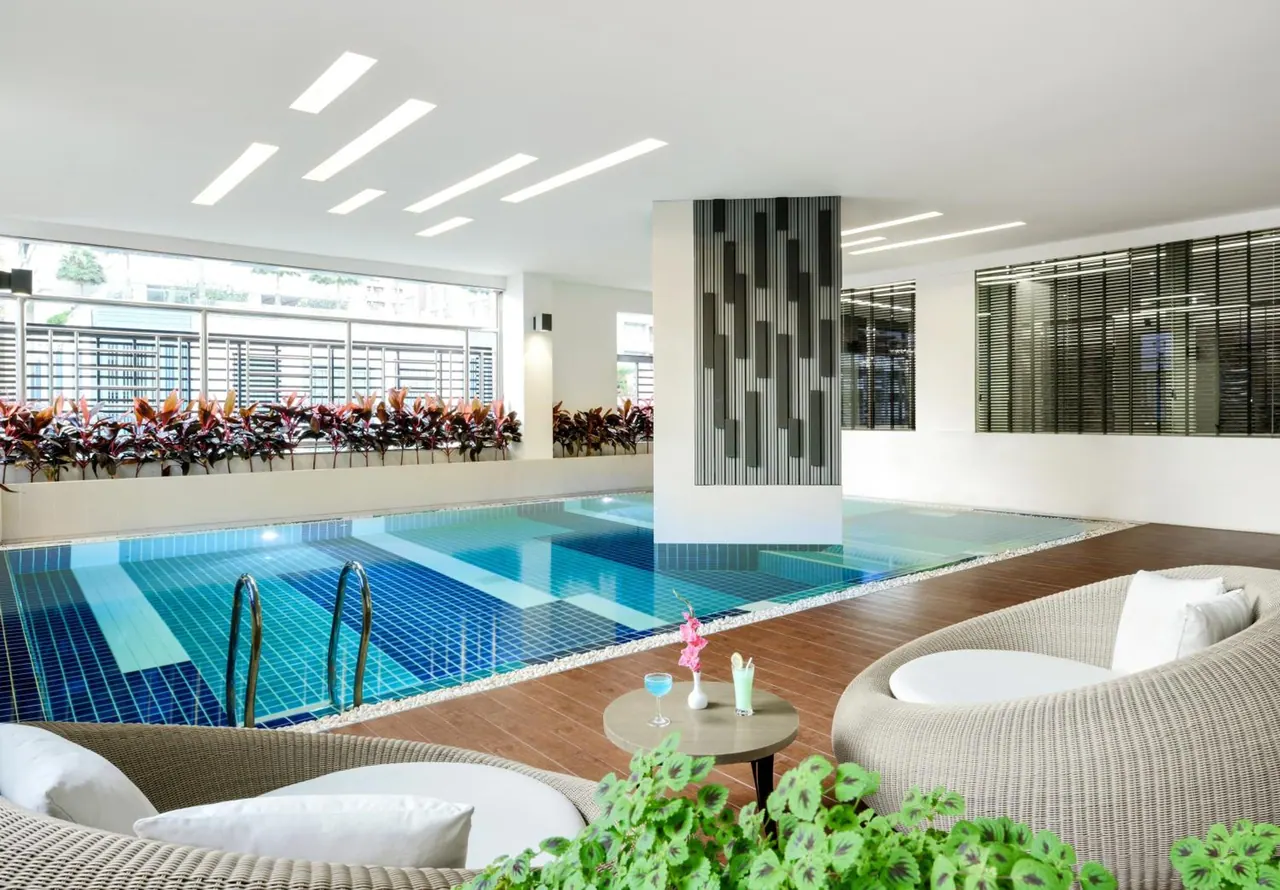 An indoor swimming pool with blue tiles, surrounded by wicker lounge seating and a small round table with drinks.