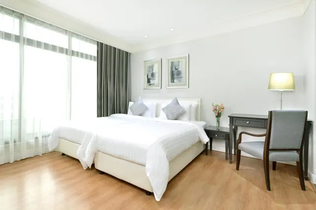A bedroom with a made double bed, a dark wooden desk and chair, a bedside table with a flower vase, and tall windows with sheer curtains.
