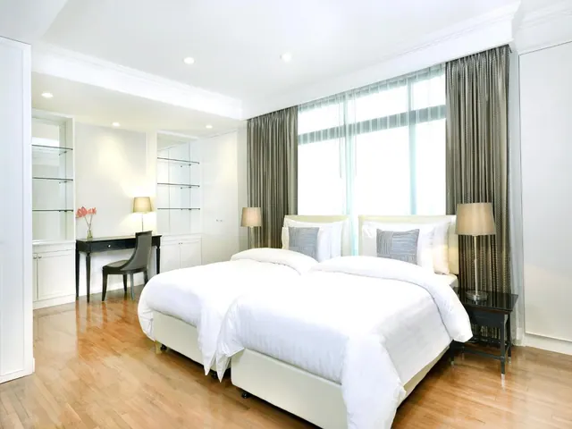 A bedroom with two beds made with white bedding, a dark wooden desk and chair, built-in white cabinets and glass shelving, and taupe curtains beside a bright window.