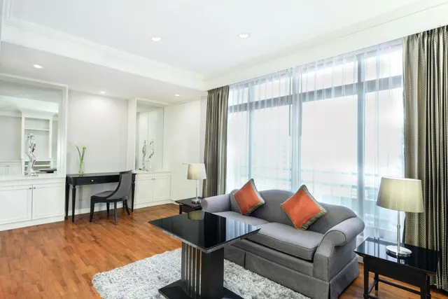 A living area with a grey sofa, two orange cushions, a black coffee table and a desk with a chair, beside floor-to-ceiling windows with sheer curtains.