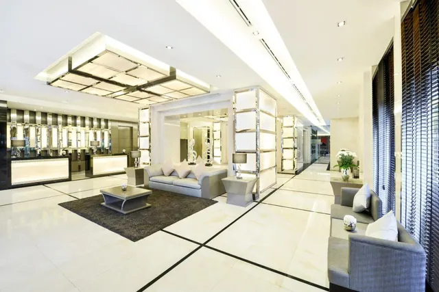 A bright lobby with a reception desk on the left, grey sofas around a dark rug and coffee table, and mirrored pillars.