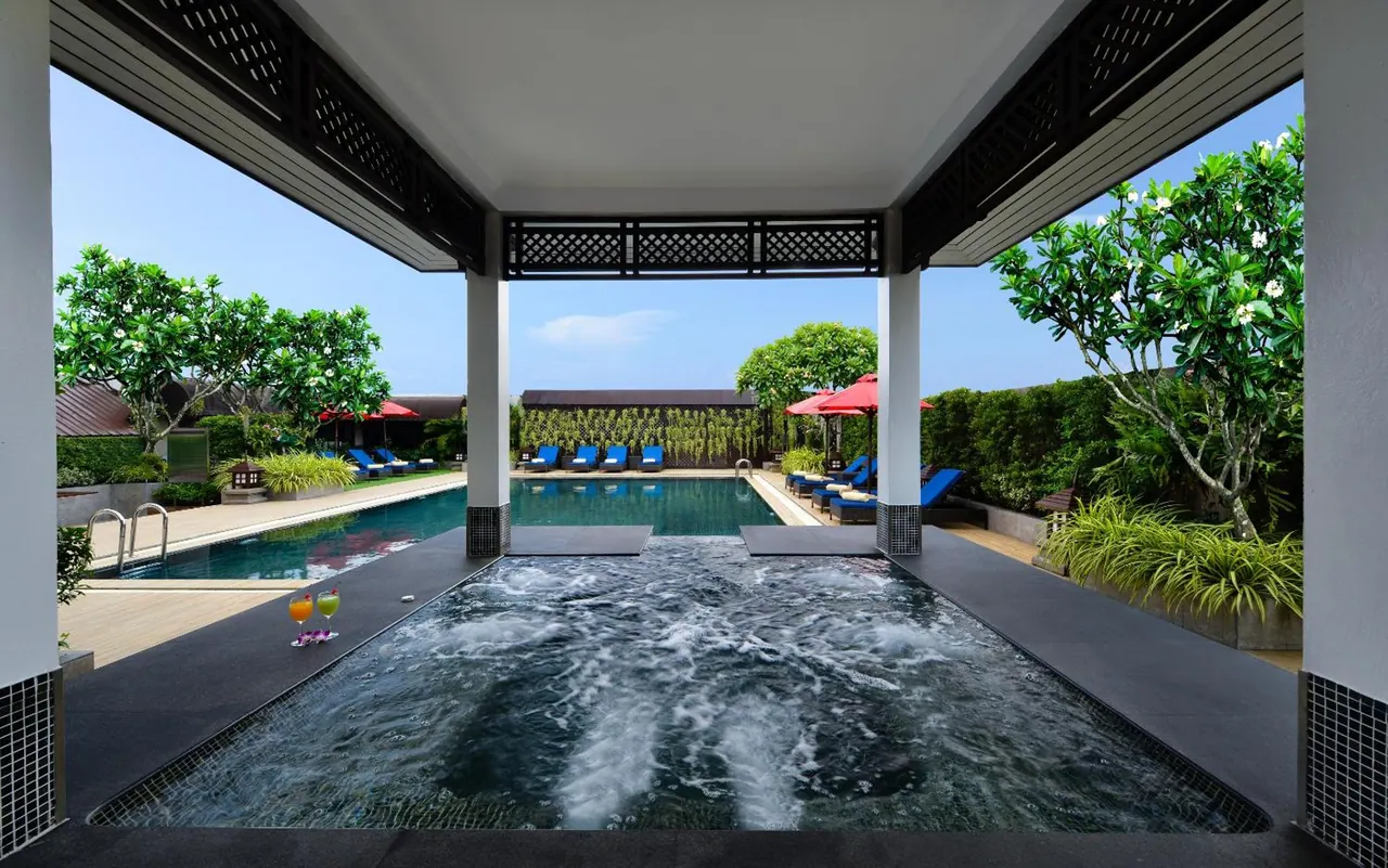 A covered pavilion frames a bubbling hot tub in the foreground, with a larger swimming pool, blue sun loungers and red parasols beyond, among frangipani trees and hedges.