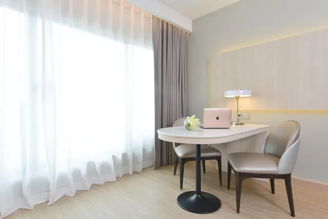 A white desk with two upholstered chairs, a laptop and a table lamp sit in a room beside sheer white curtains.