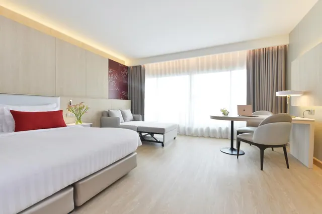 A hotel bedroom with a large white bed, a grey sofa and ottoman, a round desk with a laptop and chairs, and sheer-curtained floor-to-ceiling windows.