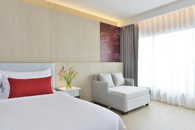 A bedroom with a made double bed with a red cushion, a grey pull-out sofa bed beside a large window with sheer white curtains, and a vase of flowers on a bedside table.