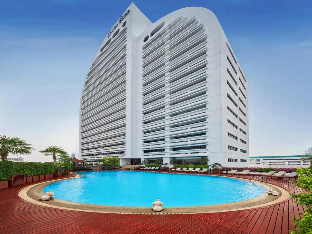 A white louvred hotel tower rises above a round blue swimming pool on a wooden deck, with sun loungers along the far edge.