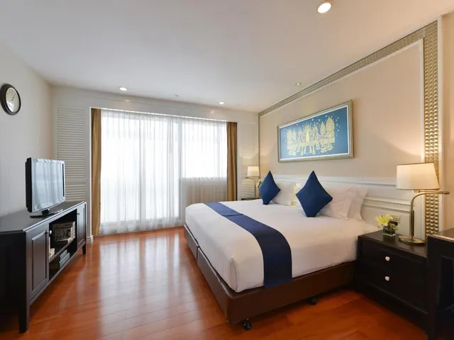 A bedroom with a king bed with blue accent pillows and a blue runner, a flat-screen TV on a dark wooden cabinet, and a window with sheer curtains.