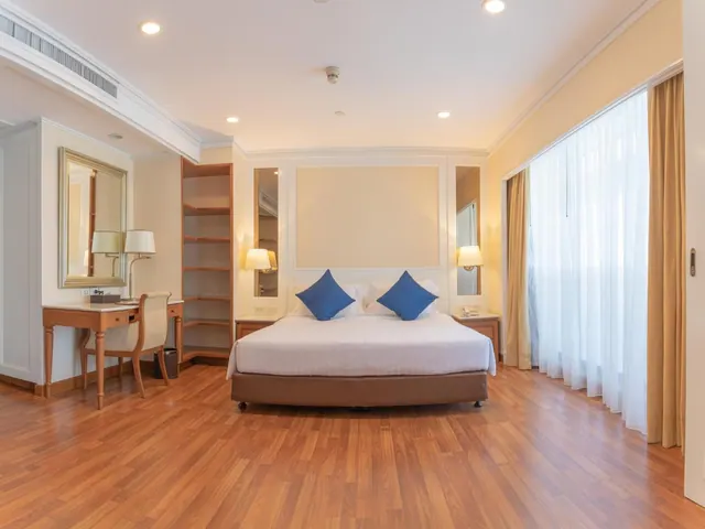 A bedroom with a large bed with blue cushions, a wooden writing desk with a chair, open wall shelving, and a tall window with sheer curtains.