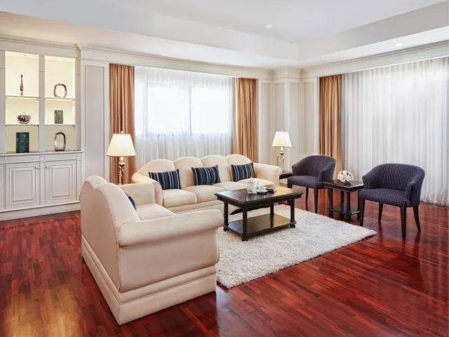 A furnished living room with a cream sofa and armchair, a dark wood coffee table on a shag rug, two striped armchairs, a built-in display cabinet and sheer-curtained windows.
