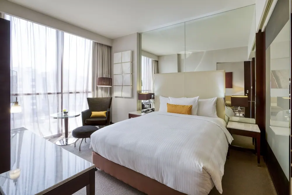 A hotel bedroom with a king bed, an armchair and round side table beside floor-to-ceiling windows, a glass desk in the foreground and a mirrored wardrobe on the right.