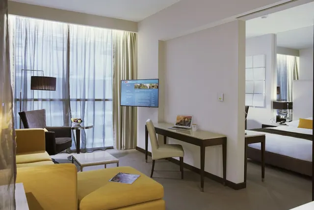 A hotel suite living area with yellow sofas, a coffee table, a desk with a laptop and wall-mounted television, and a bed visible through an opening on the right.