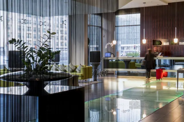 A hotel lobby with a reception desk, green lounge seating, a potted plant and a glass-floored walkway, with a blurred person pulling a red suitcase.