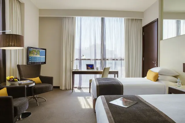 A hotel bedroom with two single beds, an armchair and seating area, a desk by the window and a wall-mounted television.