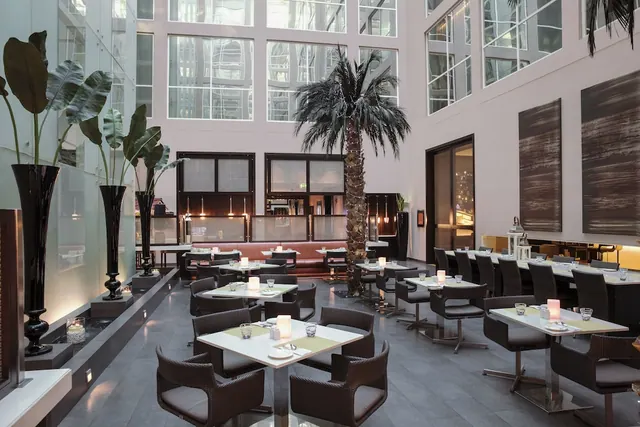A restaurant with rows of tables and woven chairs beneath a tall atrium, with a palm tree, large potted plants and a bar along the right.