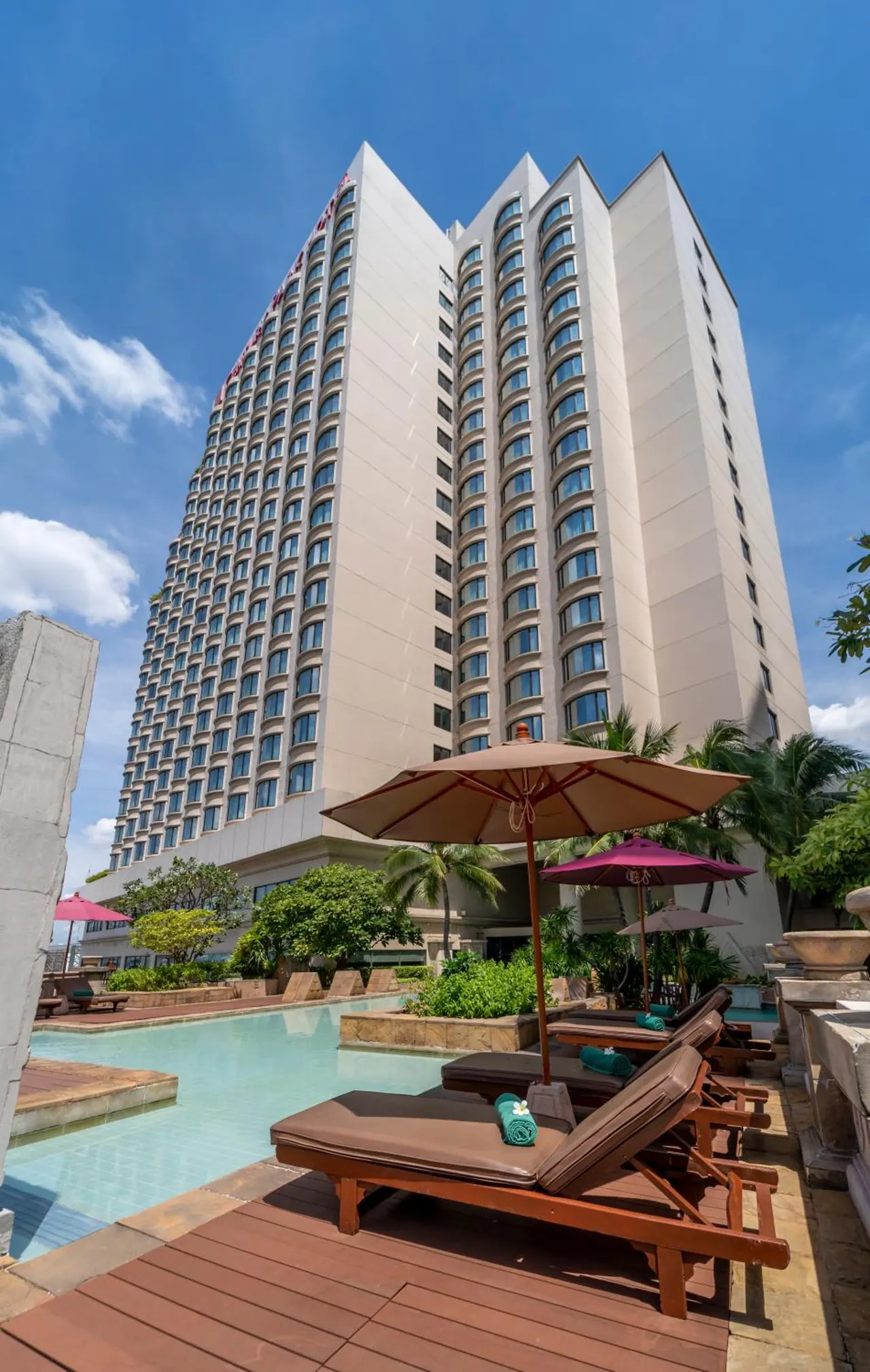 A tall hotel tower rises above a swimming pool, with sun loungers and parasols on the wooden deck under a blue sky.