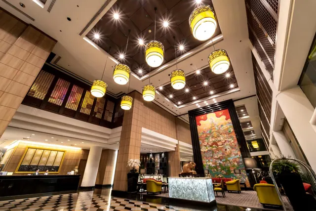 A hotel lobby with yellow pendant lamps under a coffered ceiling, a reception desk with two staff on the left, a large painting on the right and yellow lounge chairs in the seating area.