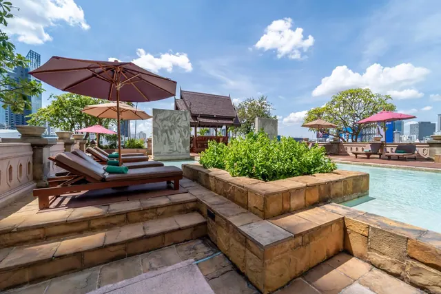 A rooftop pool with sun loungers and shade umbrellas, a planted section in the pool, a Thai-style pavilion, and city buildings in the background under a blue sky.