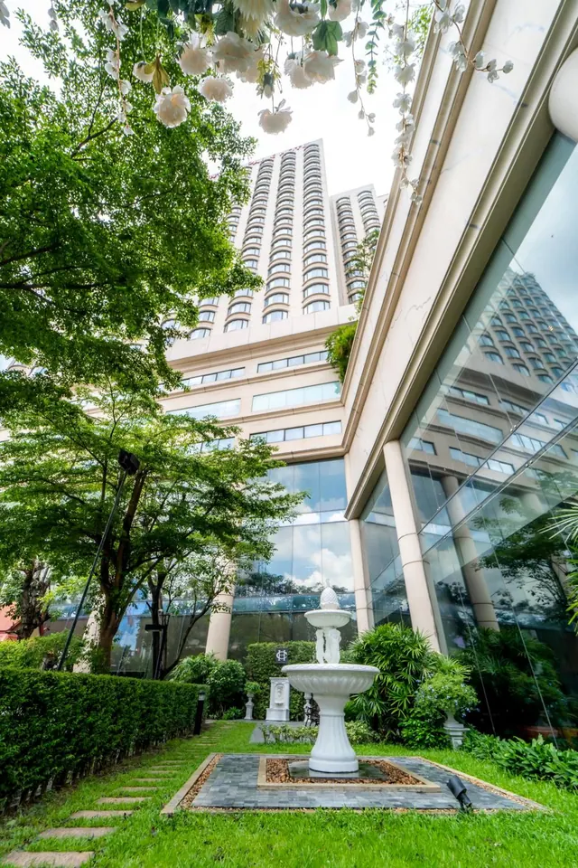 A white tiered fountain stands in a paved garden court beside a glass-fronted hotel tower, with trees and a stepping-stone path across the lawn.