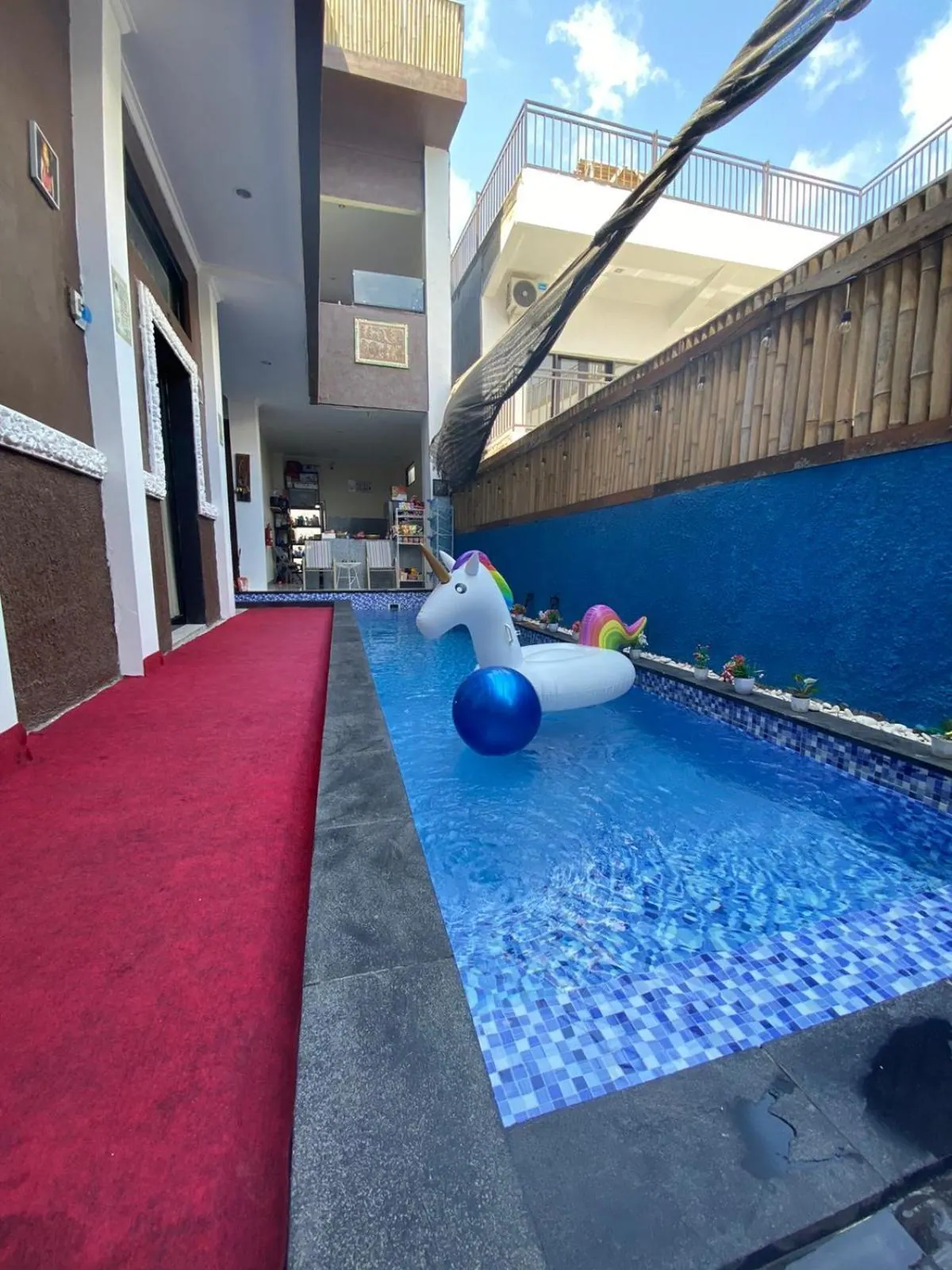 A narrow swimming pool with blue mosaic tiles holds a unicorn pool float and a blue ball, beside a red carpeted walkway and a multi-storey building.