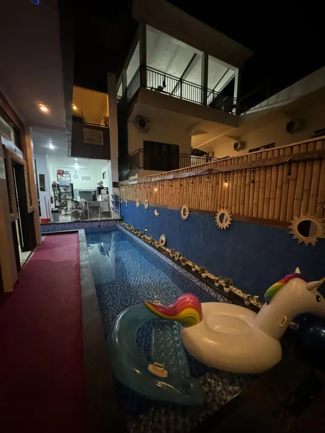 A narrow tiled swimming pool at night with a unicorn inflatable and a blue inflatable floating in it, beside a bamboo fence and a two-storey building.