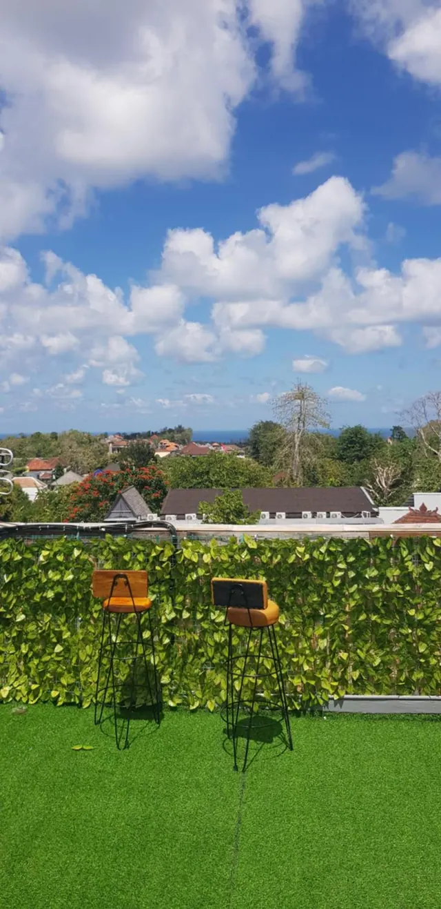 A rooftop terrace with artificial grass, a leafy green screen and two wooden bar stools, with village rooftops, trees and a thin strip of sea beyond.