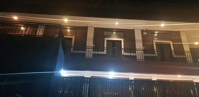 Night exterior of a two-storey building with ceiling lights on, a railed upper balcony with doors, and a dark slatted fence in the foreground.